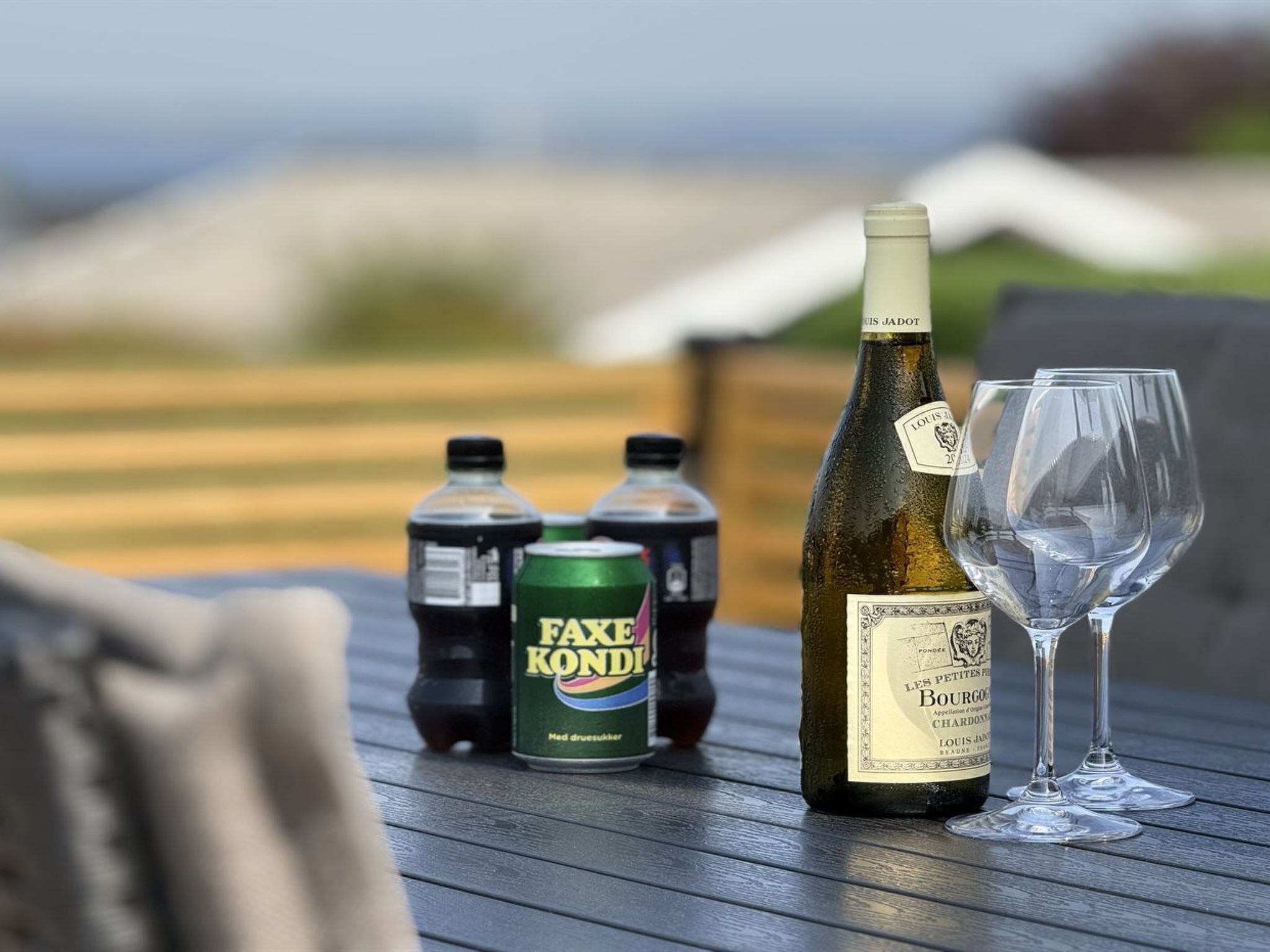 A bottle of wine, two cans of soda, and two empty wine glasses on a wooden table with a blurred outdoor background.