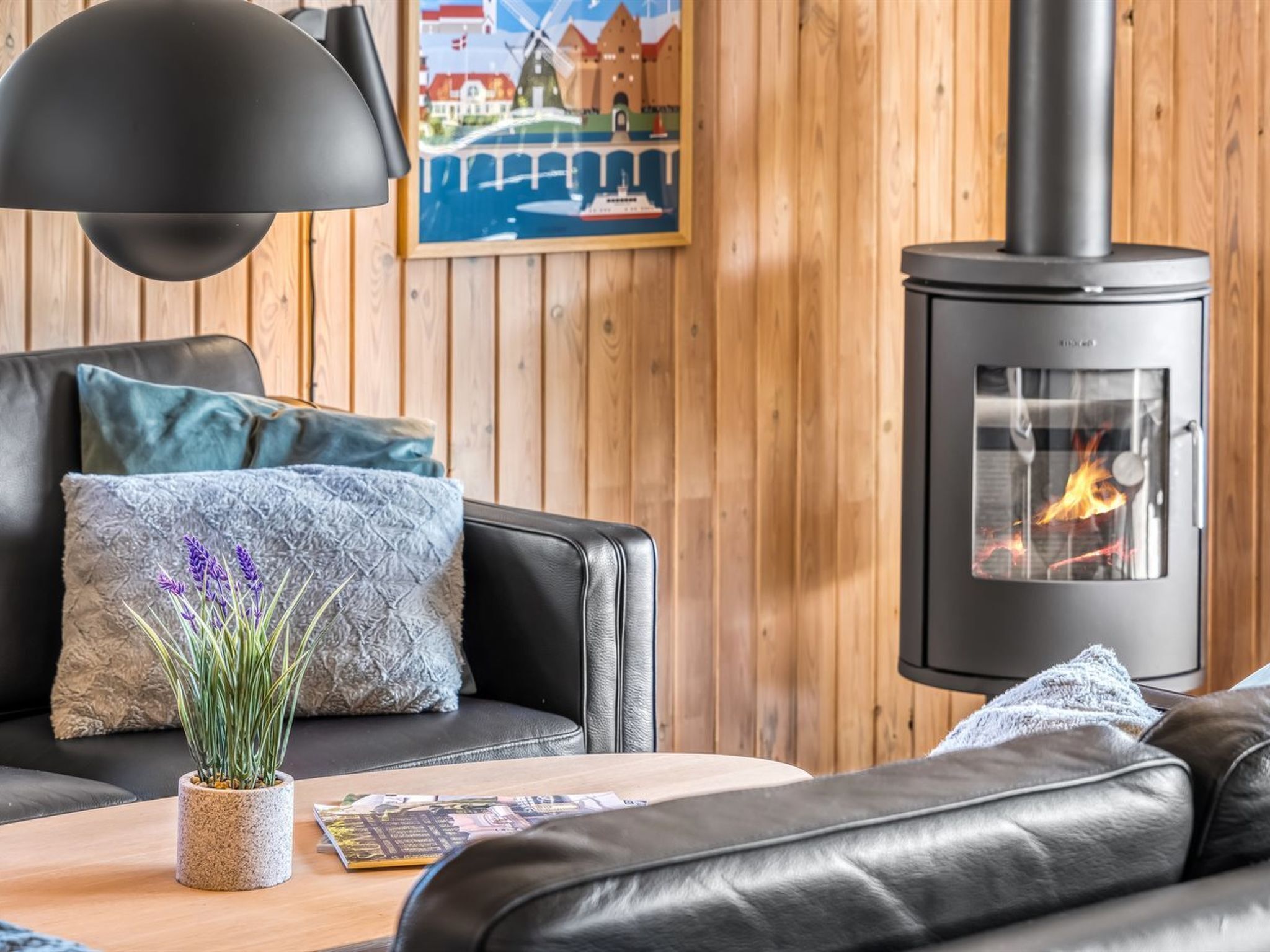 A cozy living room with a black leather sofa, a wooden coffee table, and a black pendant lamp. A framed picture of a windmill is on the wall, and a wood-burning stove is visible.