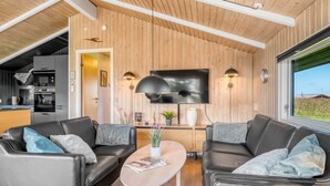 Een moderne woonkamer met houten plafond, een flatscreen televisie, twee leren banken, een salontafel en uitzicht op de natuur door een groot raam.