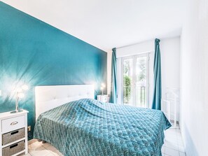 Ein Schlafzimmer mit weißem Kopfteil, einem gemusterten Bettüberwurf, einem Nachttisch mit Lampe und Blick ins Freie durch eine Glastür.