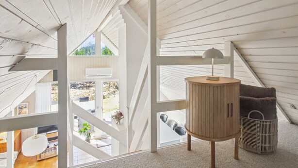 Una habitación con techo alto, vigas de madera blanca, un armario redondo de madera con una lámpara, una cesta y vistas a través de un ventanal grande.