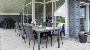 An outdoor patio with a table and chairs, a grill, and a view of a house through large windows.