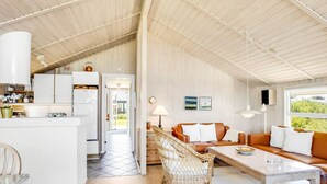 Un'ampia zona giorno con soffitto alto, un tavolo da pranzo, un divano e una sedia in vimini. Si intravede in secondo piano una cucina con mobili bianchi e un frigorifero.