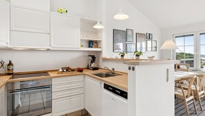 Een moderne keuken met witte kasten, een inbouwoven, een vaatwasser en een houten aanrecht. Er zijn hangende pendellampen, een raam met uitzicht en een kunstwerk in een lijst aan de muur.