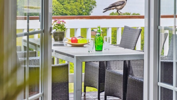 Een balkon met rieten stoelen, een tafel met een schaal fruit, een fles en een glas. Een vogel zit op de reling.