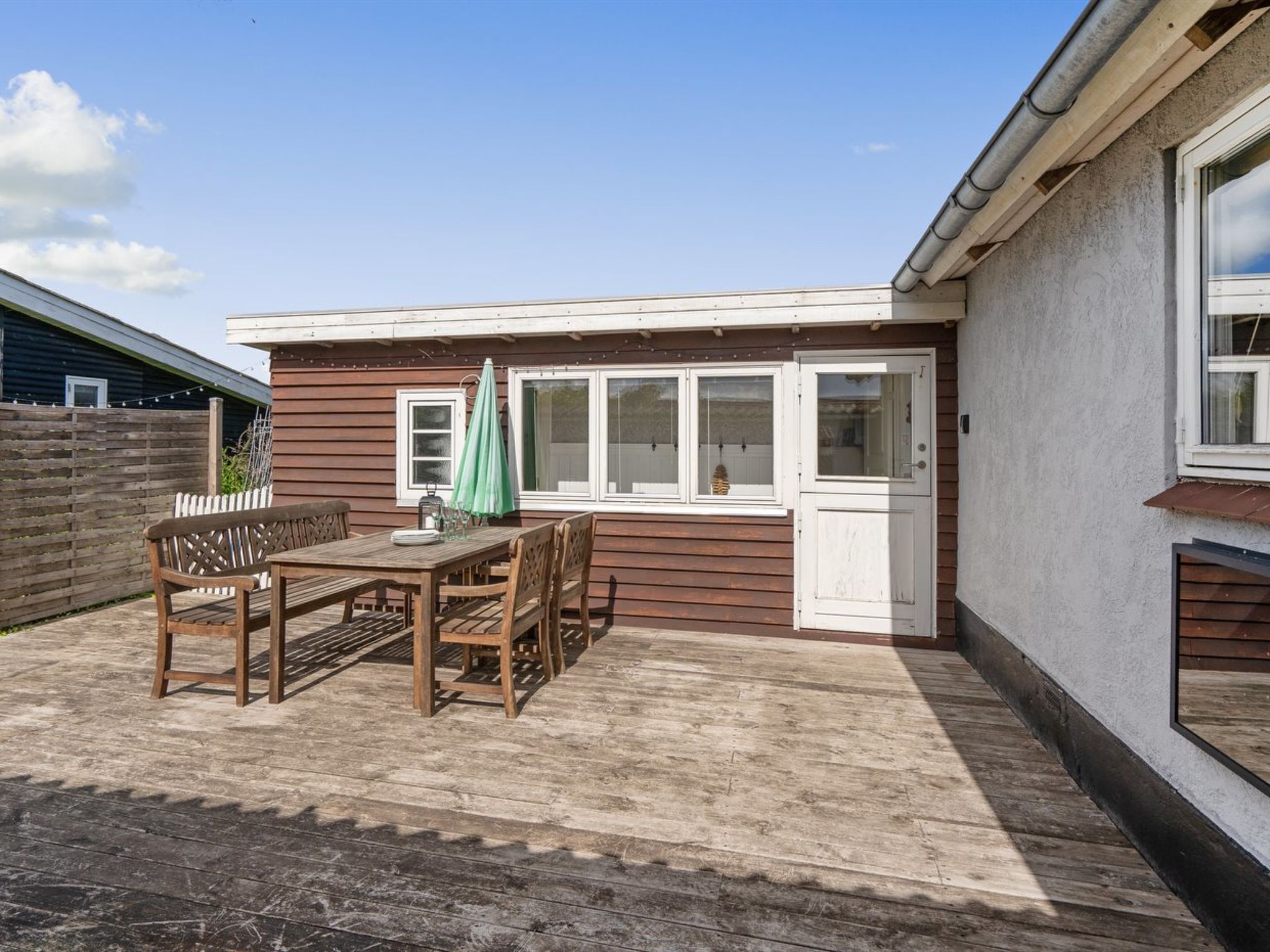 Una terraza de madera con una mesa, sillas, un paraguas verde y una puerta blanca.