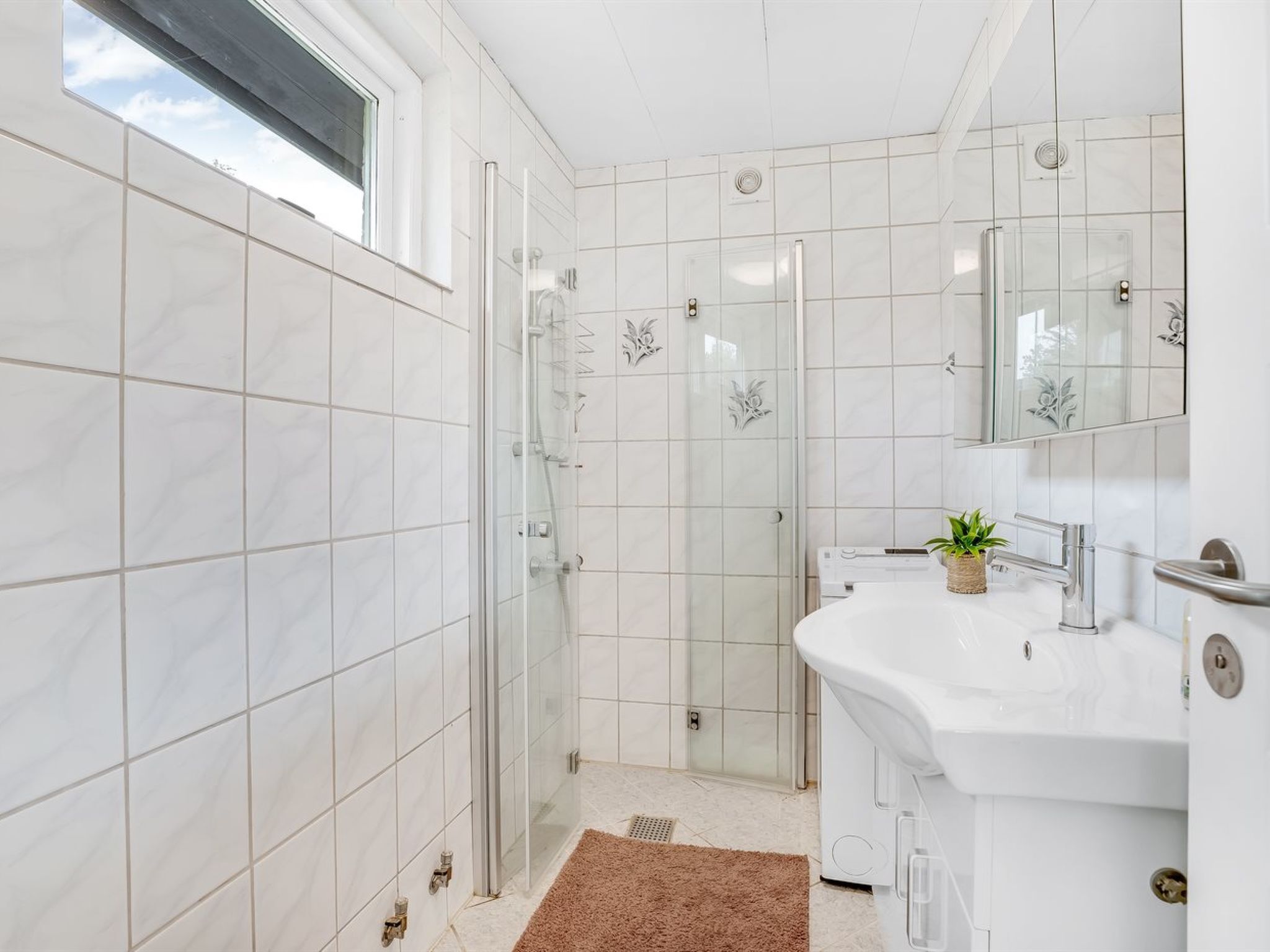 Ett modernt badrum med vita kakelplattor, en dusch med glasväggar, en vit tvättställskommod med en växt och en stor spegel.