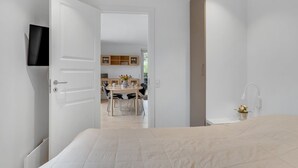 A bedroom with a bed, a TV mounted on the wall, and an open door leading to a kitchen area with a dining table and chairs.
