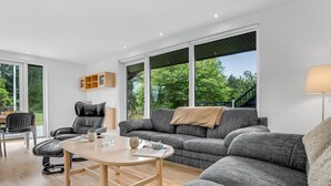 A modern living room with a grey sofa, a wooden coffee table, and a black leather chair. There is a large window with a view of greenery outside.