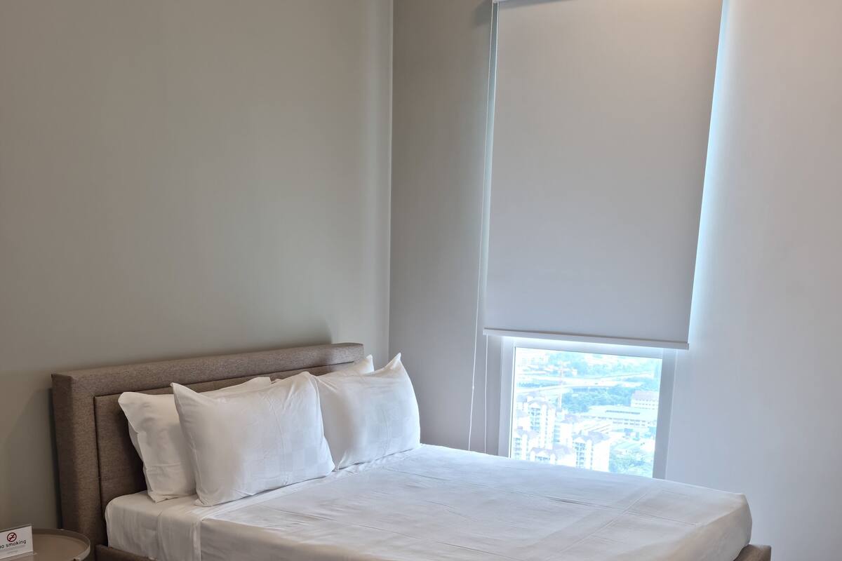 A neatly made bed with white bedding in a room with wooden flooring and a large window with a view of buildings.