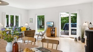 A modern living room with a sofa, chairs, a dining table with a bottle, glasses, and a magazine. There is a TV on a stand, a bookshelf, and a view of a garden through a glass door. - "Heidl" - 1.3km to the fjord by Interhome (Rørvig)
