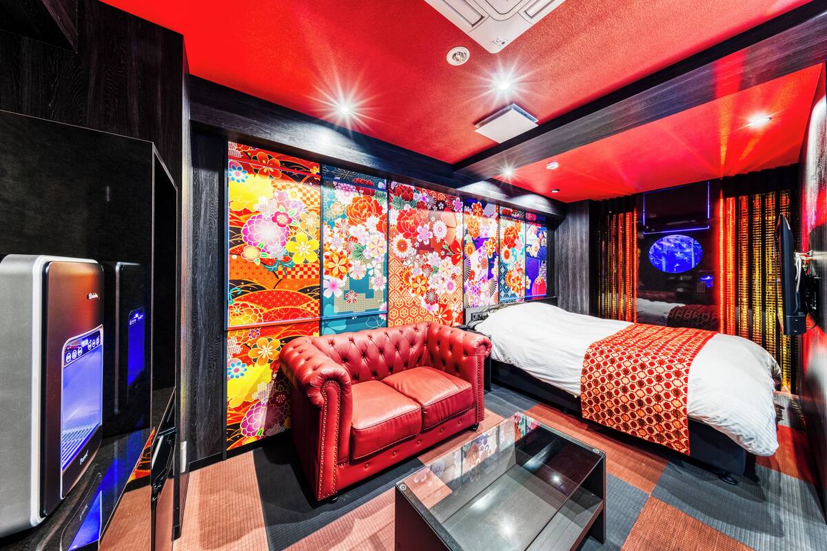 Una habitación de hotel moderna con un sofá de cuero rojo, una cama con manta estampada y una pantalla digital grande en la pared.