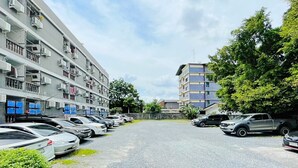 Un parking rempli de voitures, un immeuble doté de balcons et une rue bordée d’arbres.