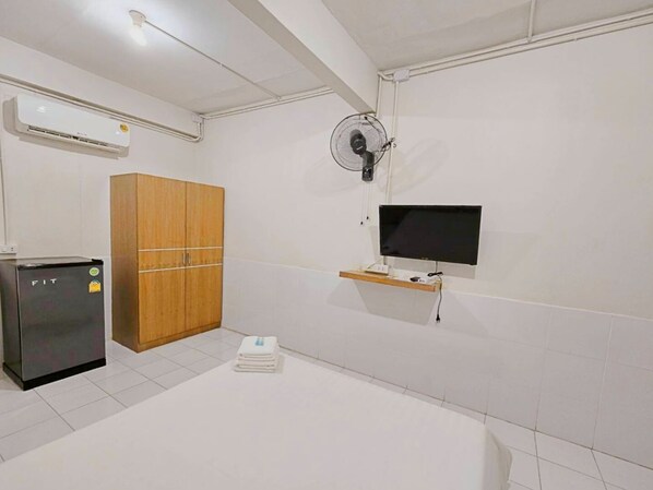 Una habitación de hotel con cama, televisor montado en la pared, armario de madera, refrigerador pequeño y ventilador de techo.