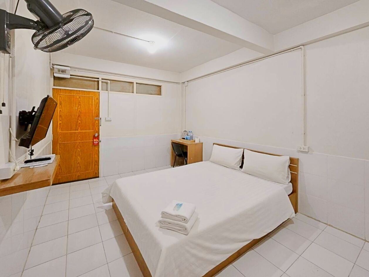 Een hotelkamer met een bed, een bureau met een computer, een stoel, een houten deur en een ventilator aan de muur.
