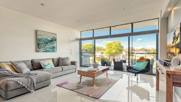 Una sala de estar moderna con un sofá gris, una mesa de centro y vista a un jardín a través de amplias ventanas.