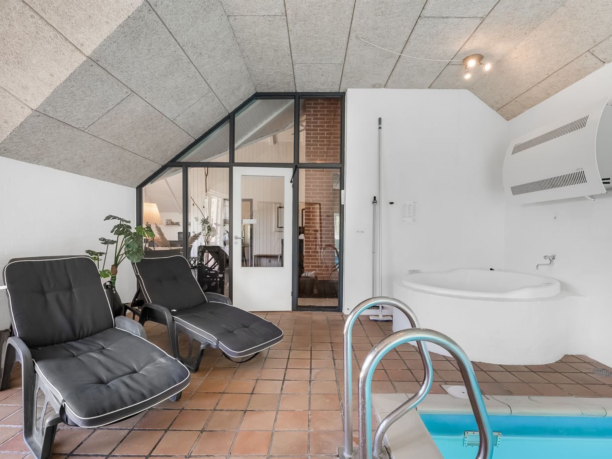 Un espace piscine intérieur moderne, doté de deux chaises longues, d’une porte vitrée donnant sur une pièce et d’une baignoire blanche.