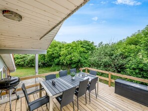 A covered outdoor deck with a table and chairs, a grill, and a view of a lush garden.