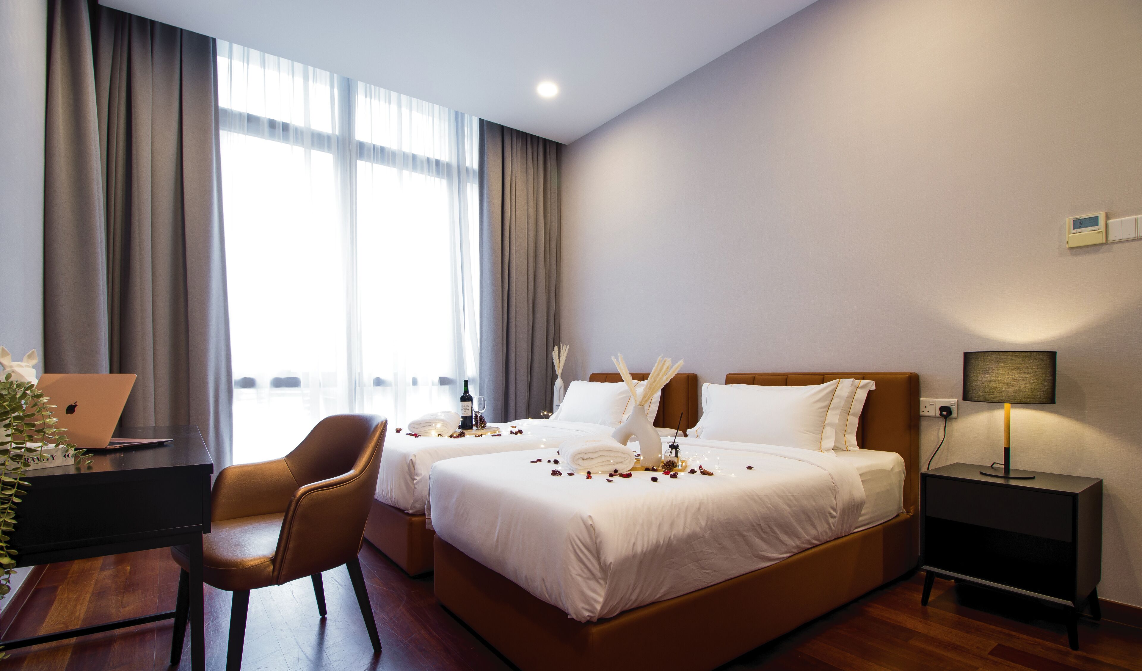 A hotel room with a bed, two chairs, a desk with a laptop, a bedside table with a lamp, and a window with curtains.