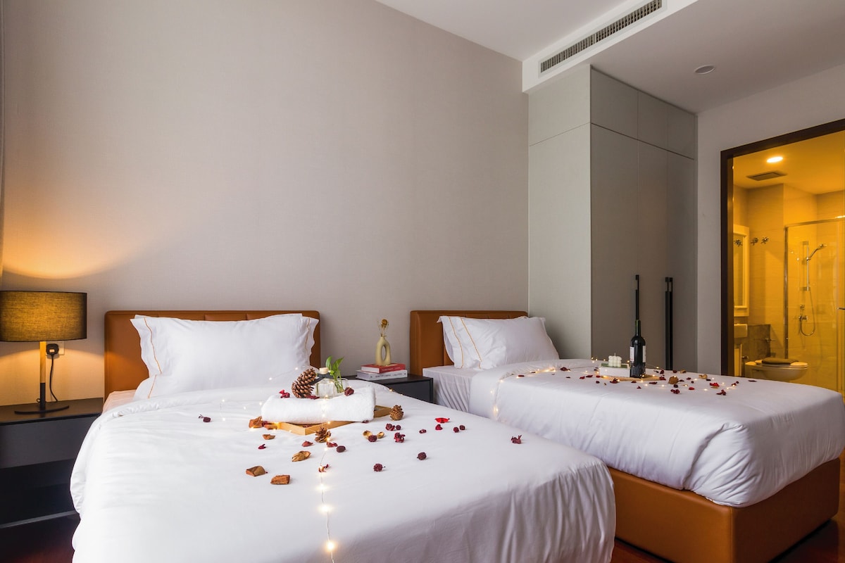 A hotel room with two beds, a bedside table with a lamp, a mirror, and a shower area.