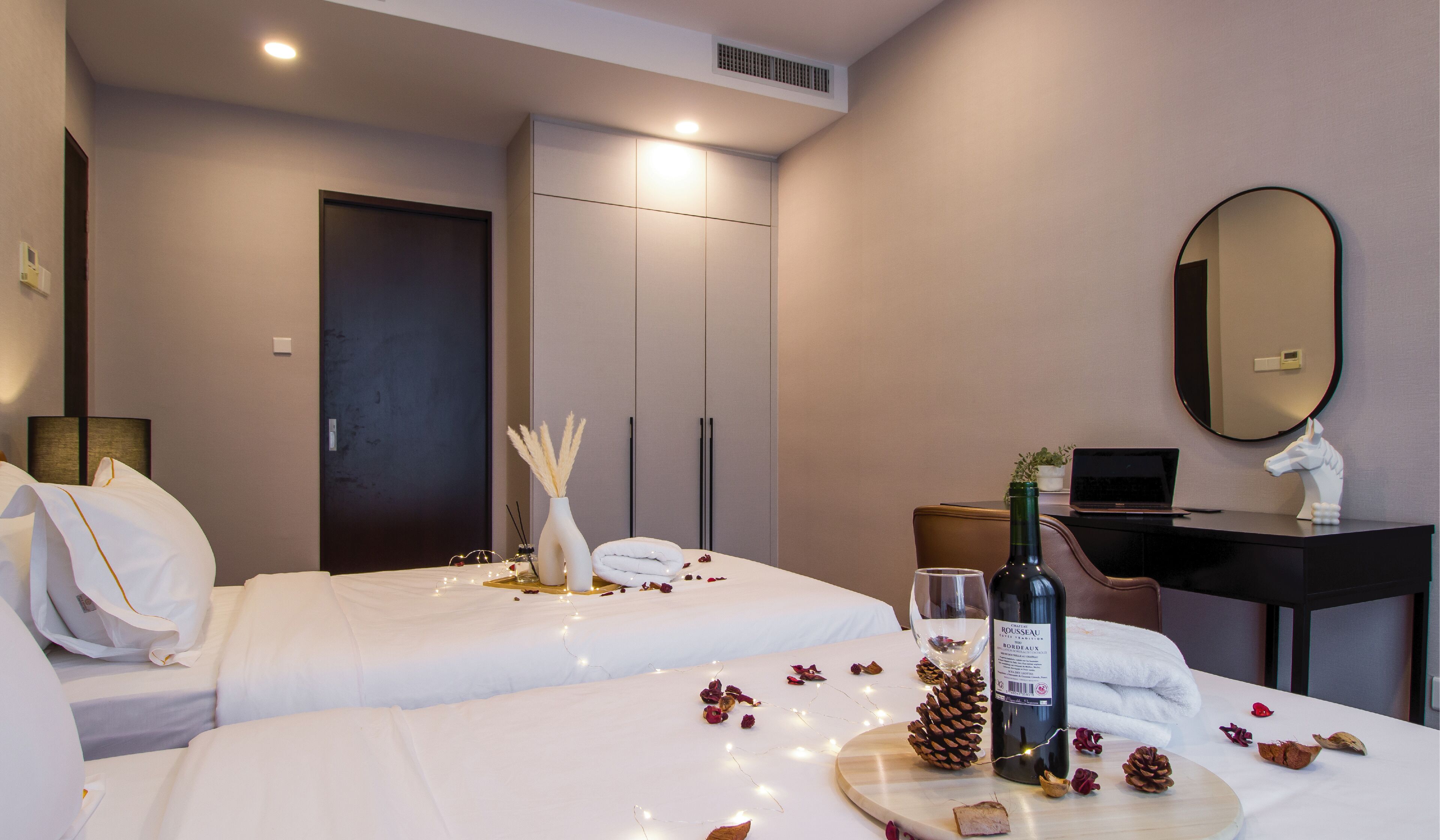 A hotel room with two beds, a desk with a laptop, a bottle of wine, and decorative items like pine cones and dried flowers.