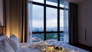Een slaapkamer met een groot raam, een bed met wit beddengoed en uitzicht op een stadsgezicht.