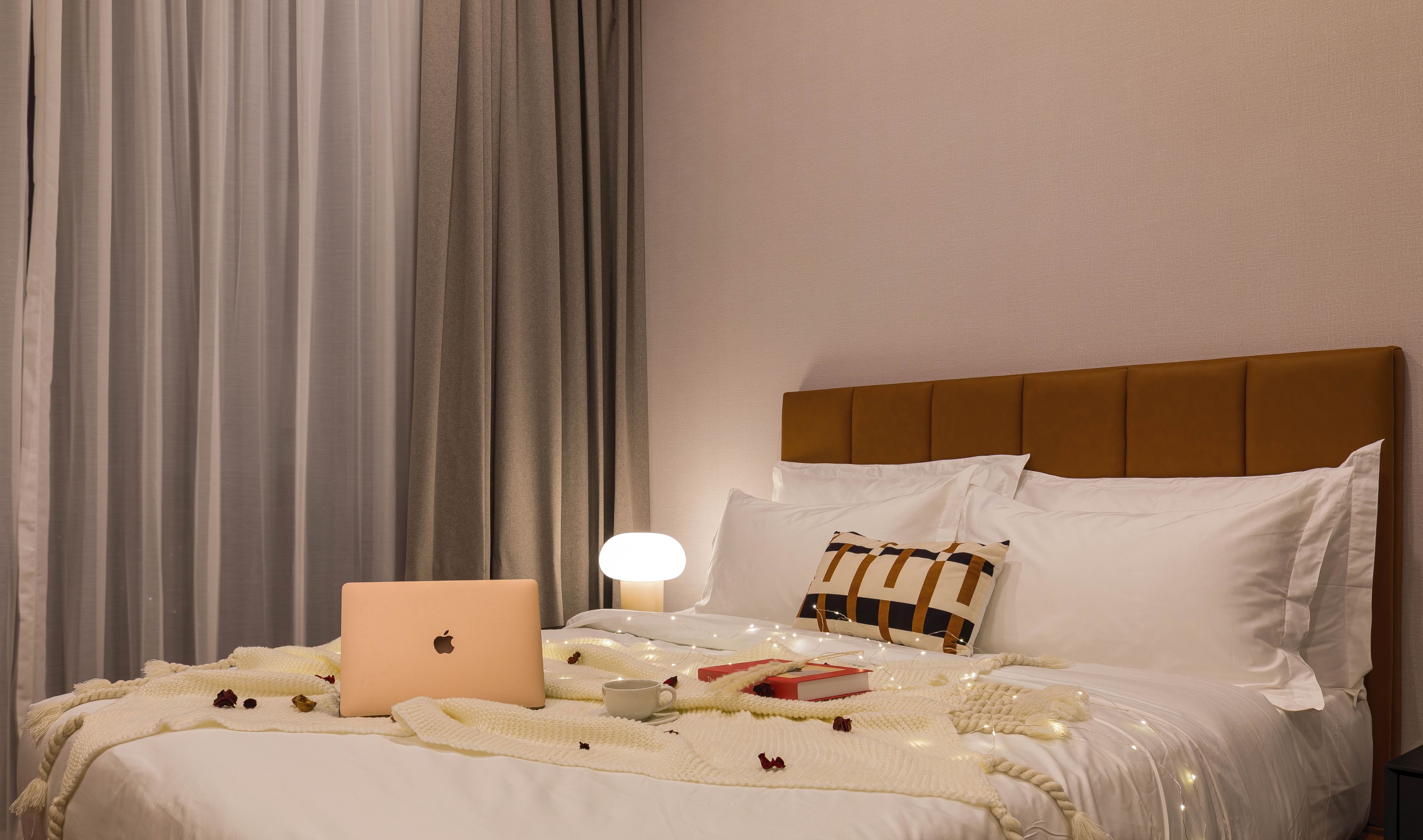 Un lit bien fait, avec un ordinateur portable, un livre et une tasse, entouré d’une couverture blanche et de guirlandes lumineuses.
