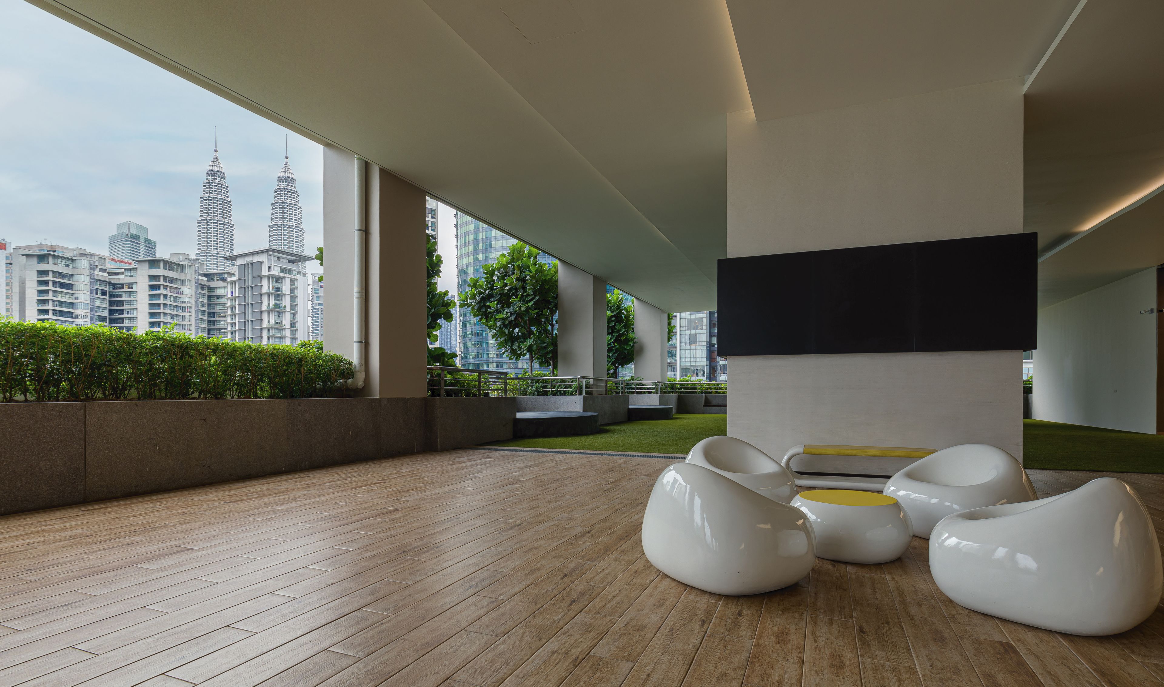 A modern outdoor area with a wooden floor, white seating, and a view of the city skyline including the Petronas Towers.