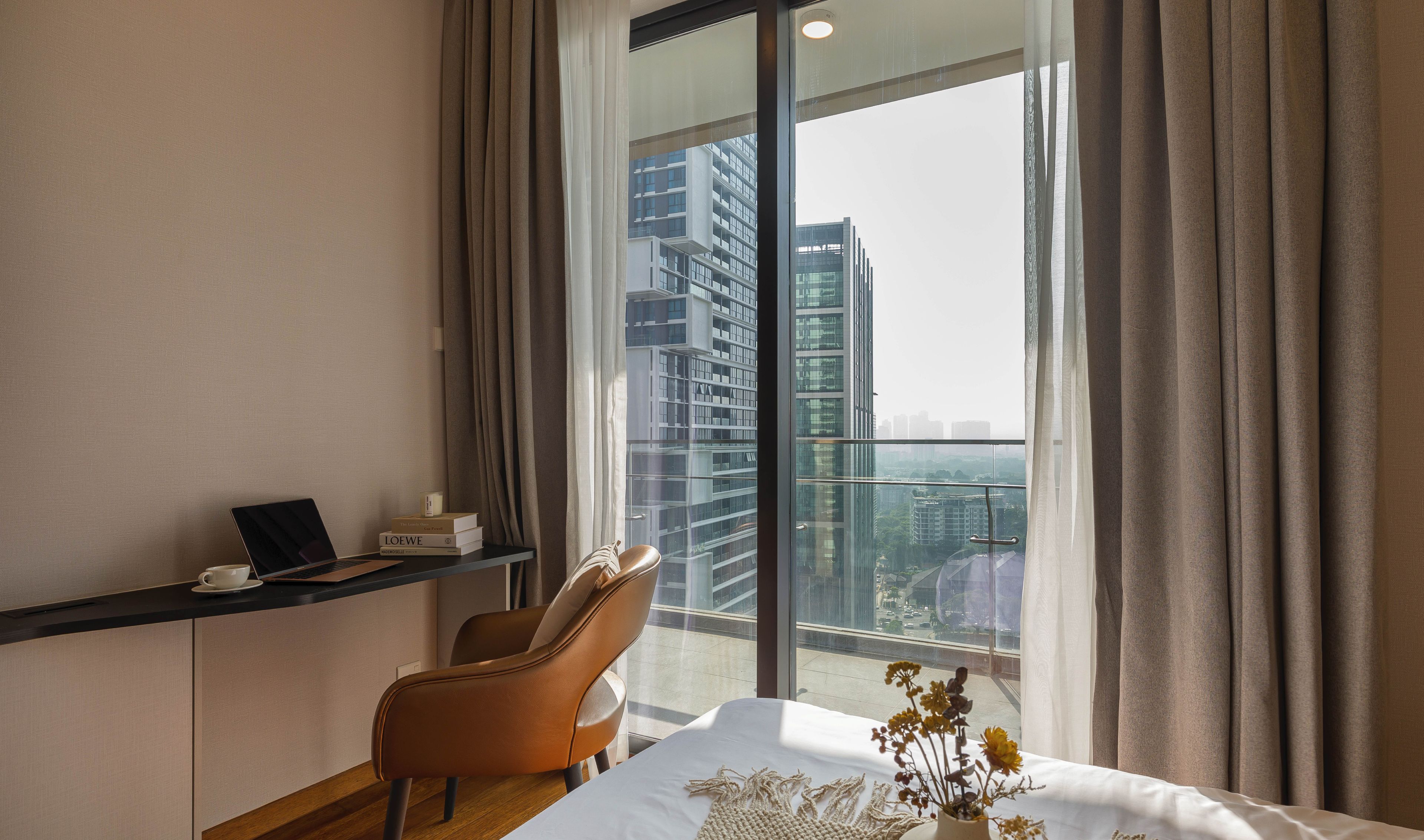 Une chambre d’hôtel moderne avec une grande fenêtre, un ordinateur portable posé sur une étagère, un fauteuil confortable et une table de chevet agrémentée d’un vase de fleurs.