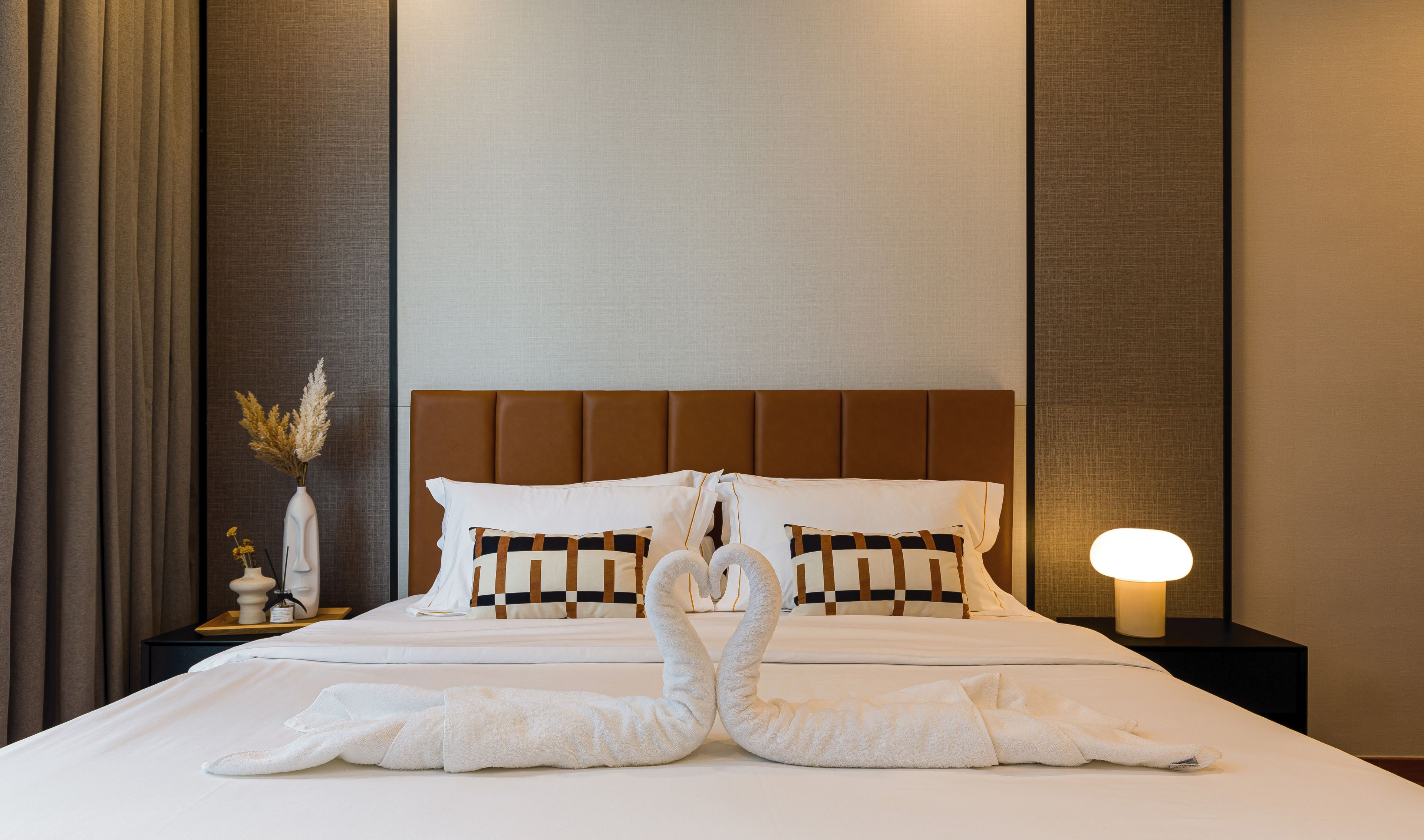 A hotel room with a neatly made bed, a bedside lamp, and a decorative vase with dried flowers.