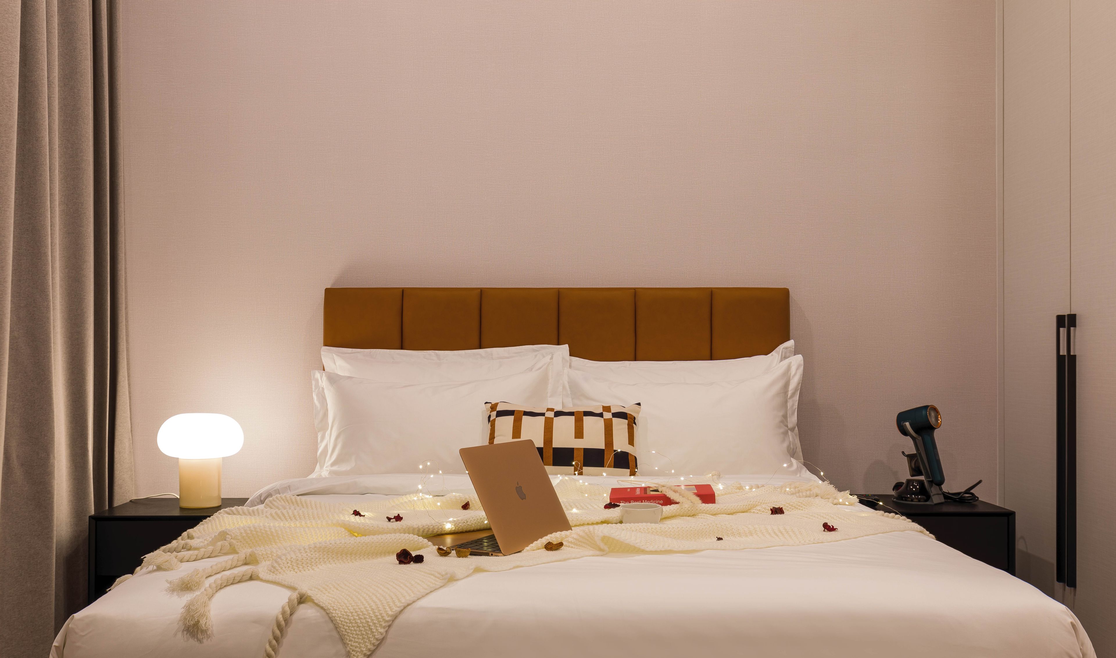 Una cama bien hecha con un ordenador portátil, una lámpara de noche y una mesita con una taza y un libro.