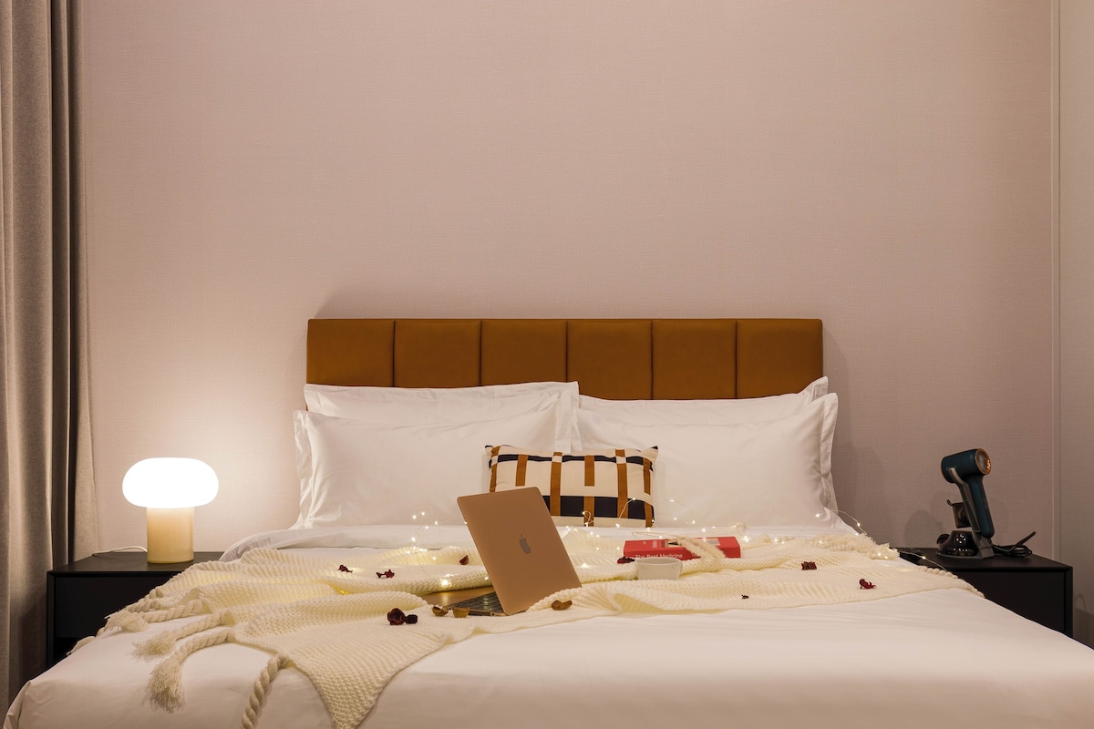 A neatly made bed with a laptop, a bedside lamp, and a small table with a cup and a book.