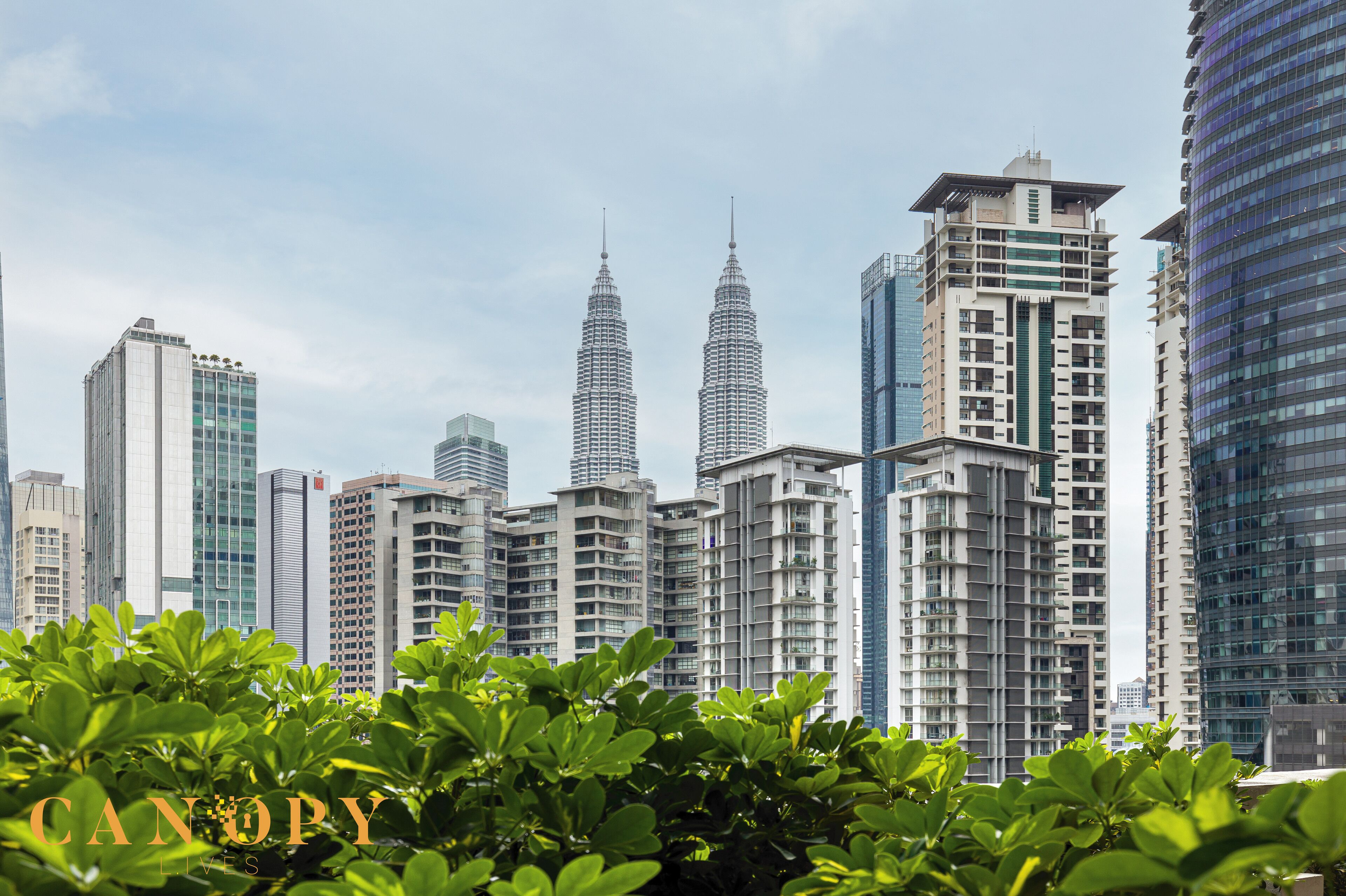 A cityscape with high-rise buildings, including the iconic Petronas Towers, and lush green foliage in the foreground.