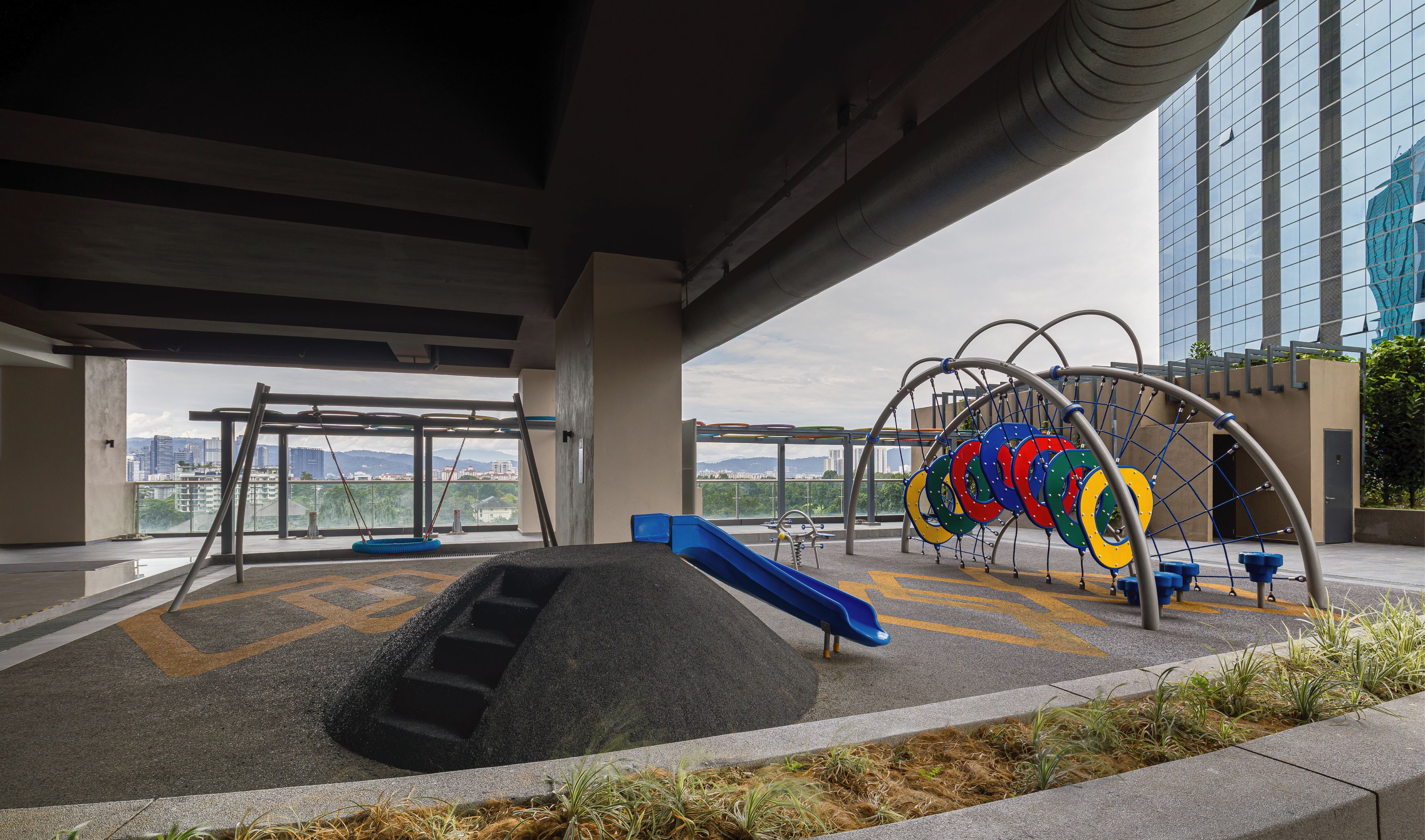An outdoor playground with slides and swings under a bridge, surrounded by a landscaped area with plants.