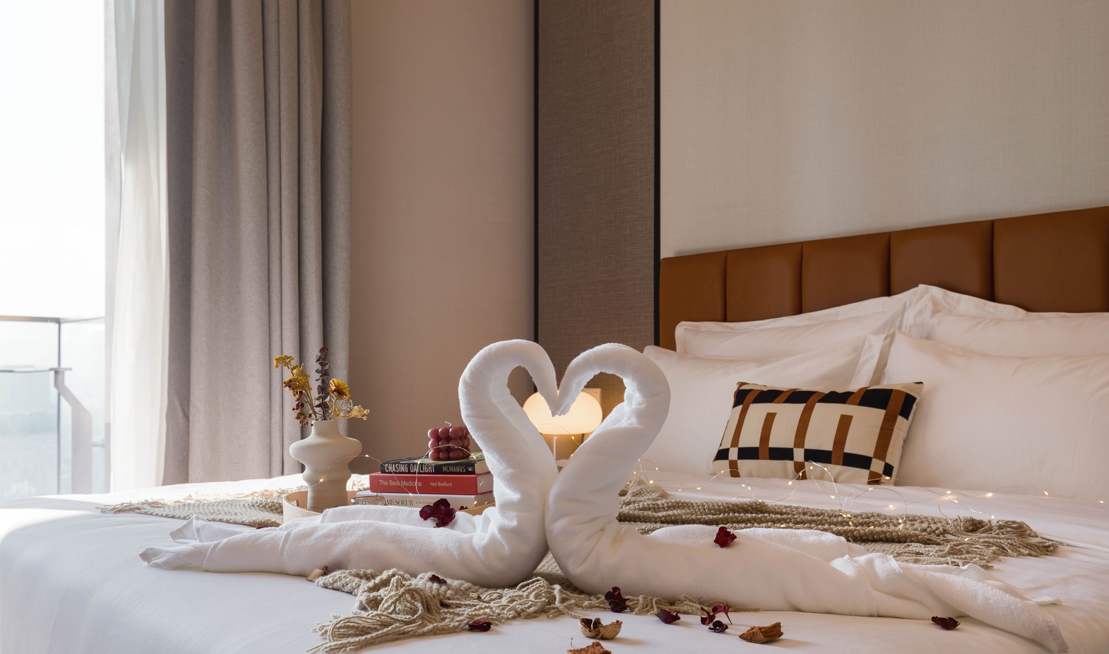 A neatly made bed with a heart-shaped towel arrangement, a bedside table with books, and a view of the outdoors.