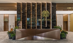 A reception area with a modern design, featuring a marble wall with vertical lines and a logo. There are potted plants and a reception desk in the foreground.