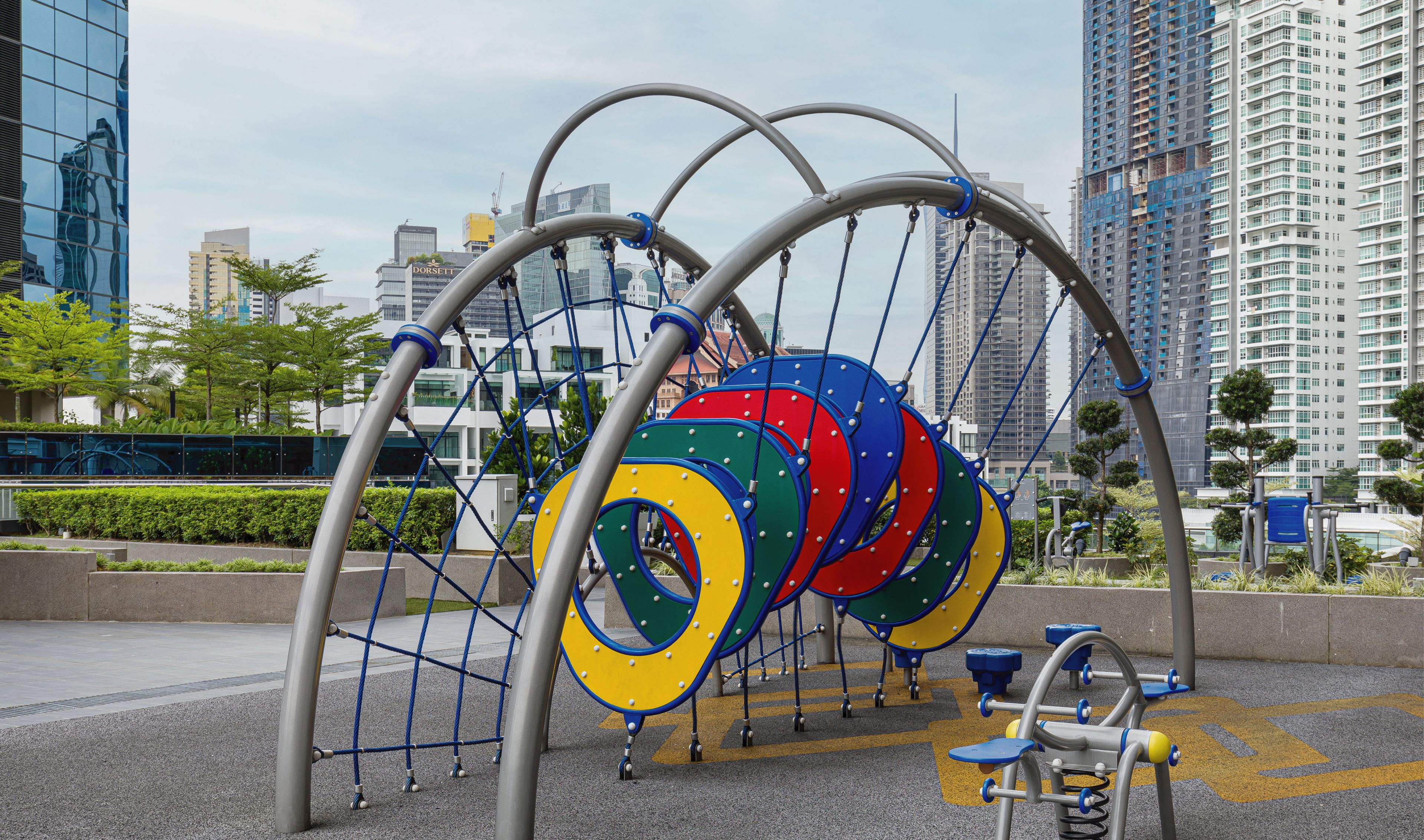 Un terrain de jeux doté d’une structure d’escalade et de balançoires, le tout entouré d’un paysage urbain.