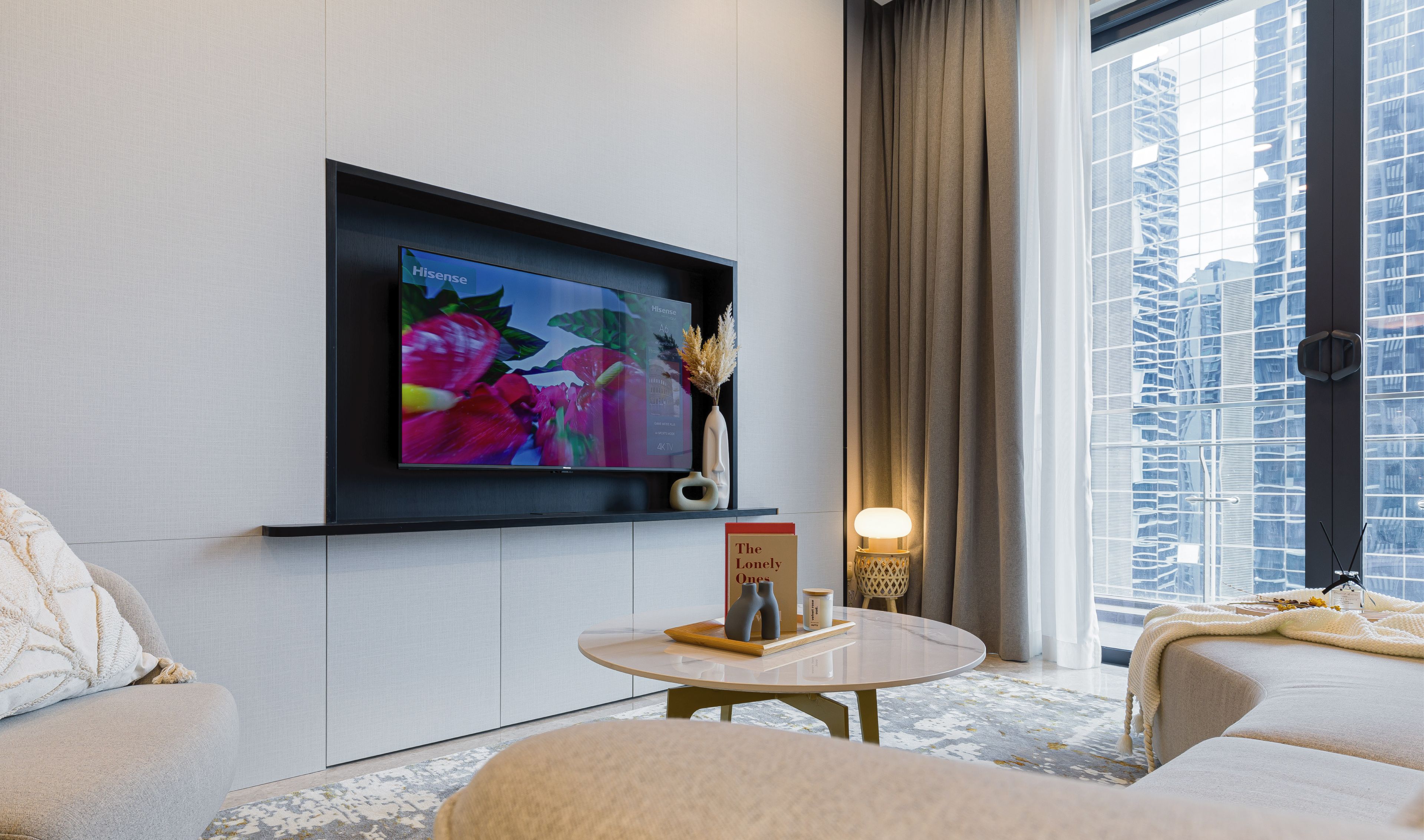 A modern hotel room with a flat-screen TV mounted on the wall, a round coffee table, a sofa, and a large window offering a city view.