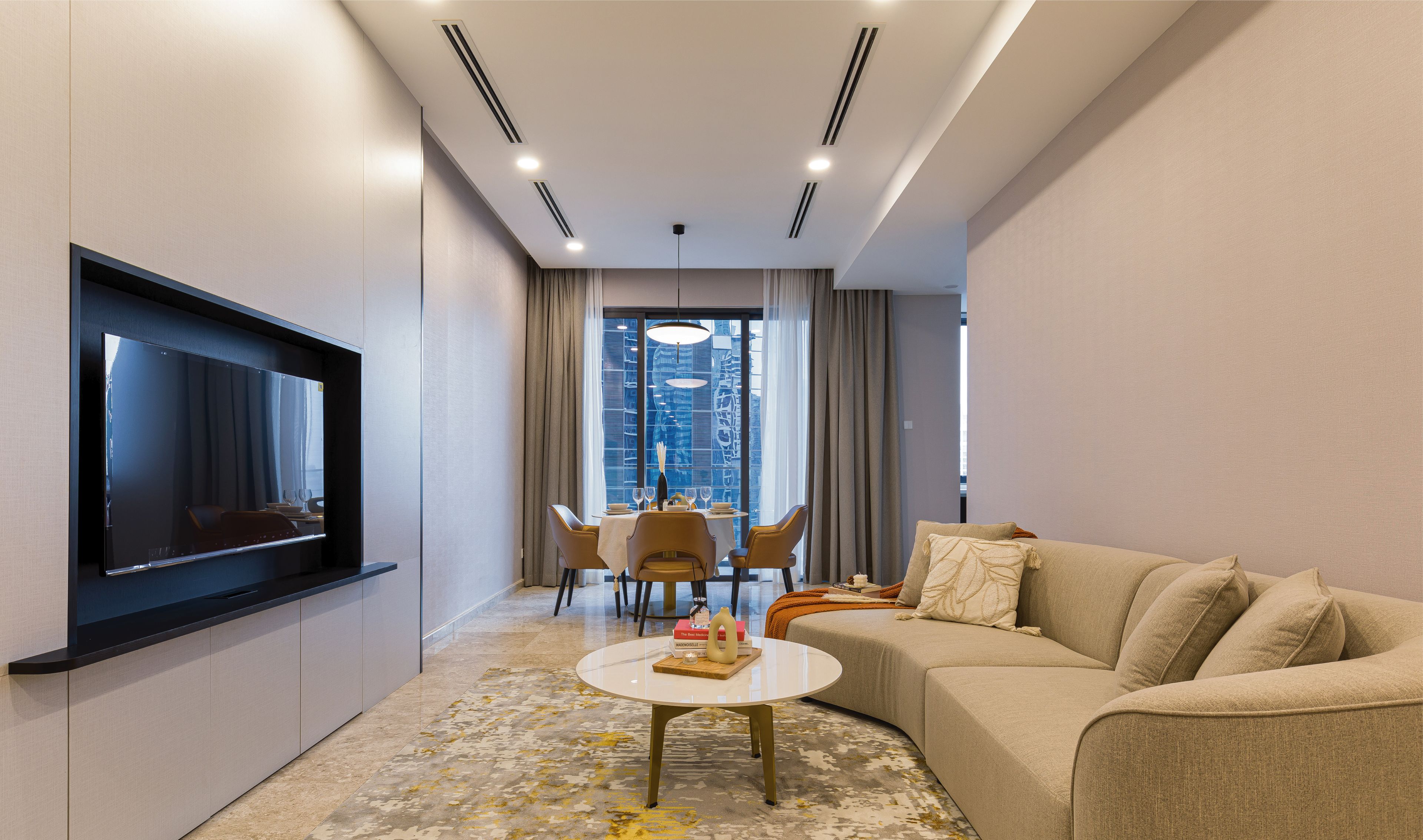 A modern hotel room with a flat-screen TV mounted on the wall, a sofa, a coffee table, and a dining area with chairs and a table.
