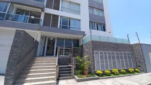 A modern building with a staircase leading to the entrance, a stone wall, and a small garden with yellow flowers. - Departamento con Vista al Mar (Lima)