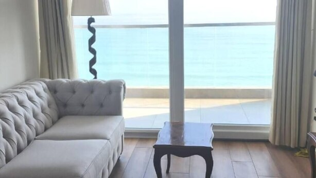 A room with a grey sofa, a small table, and a view of the sea through large windows. - Departamento con Vista al Mar (Lima)