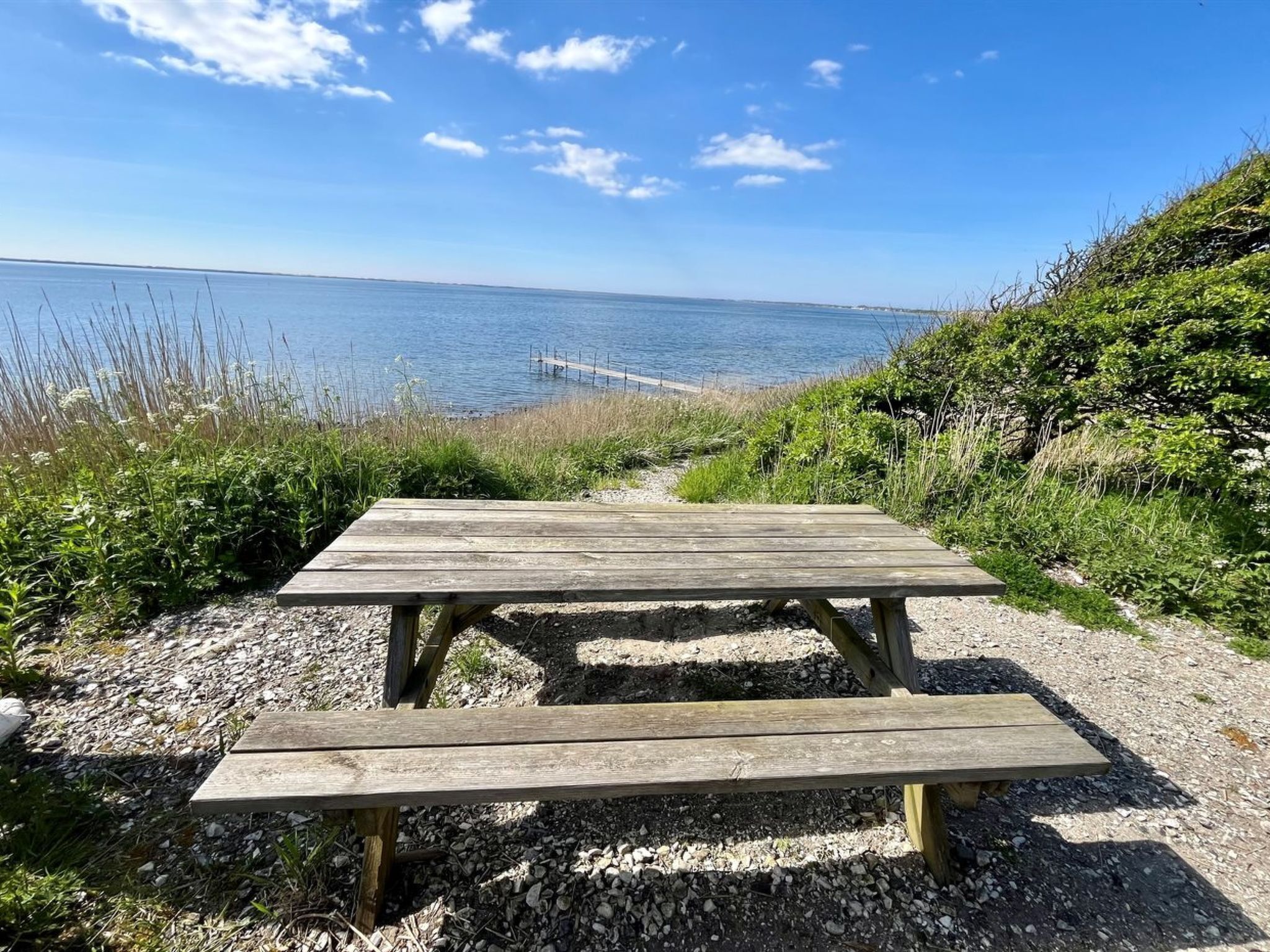 Ein Holztisch mit Bänken, umgeben von Kies und Vegetation, mit Blick auf ein Gewässer und einen Pier im Hintergrund.