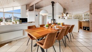A modern kitchen with a wooden ceiling, a dining area with a wooden table and leather chairs, and a large window offering a view of the outdoors.