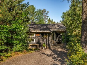 Une cabane rustique aux murs en bois, dotée d’un porche avec des sièges et d’un chemin de gravier menant à l’entrée.