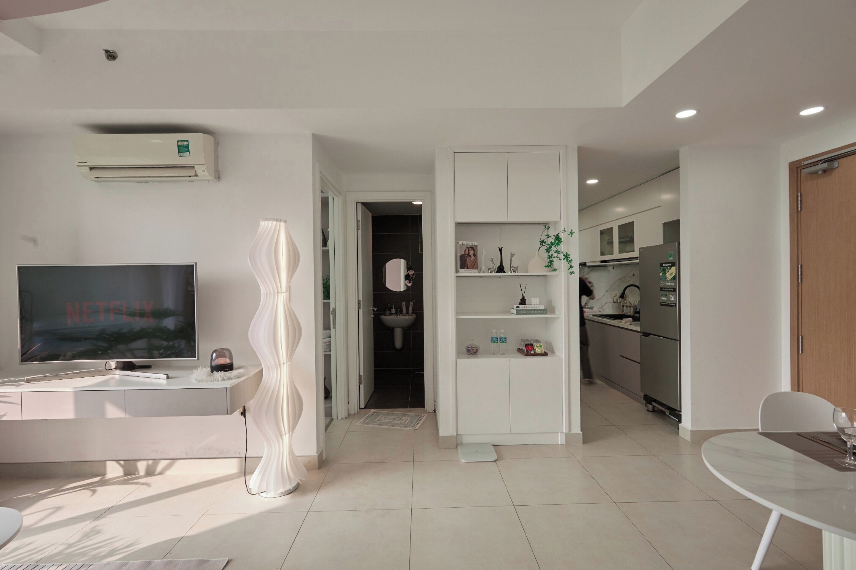 A modern living room with a flat-screen TV, a white sofa, a dining table, and a kitchen in the background.