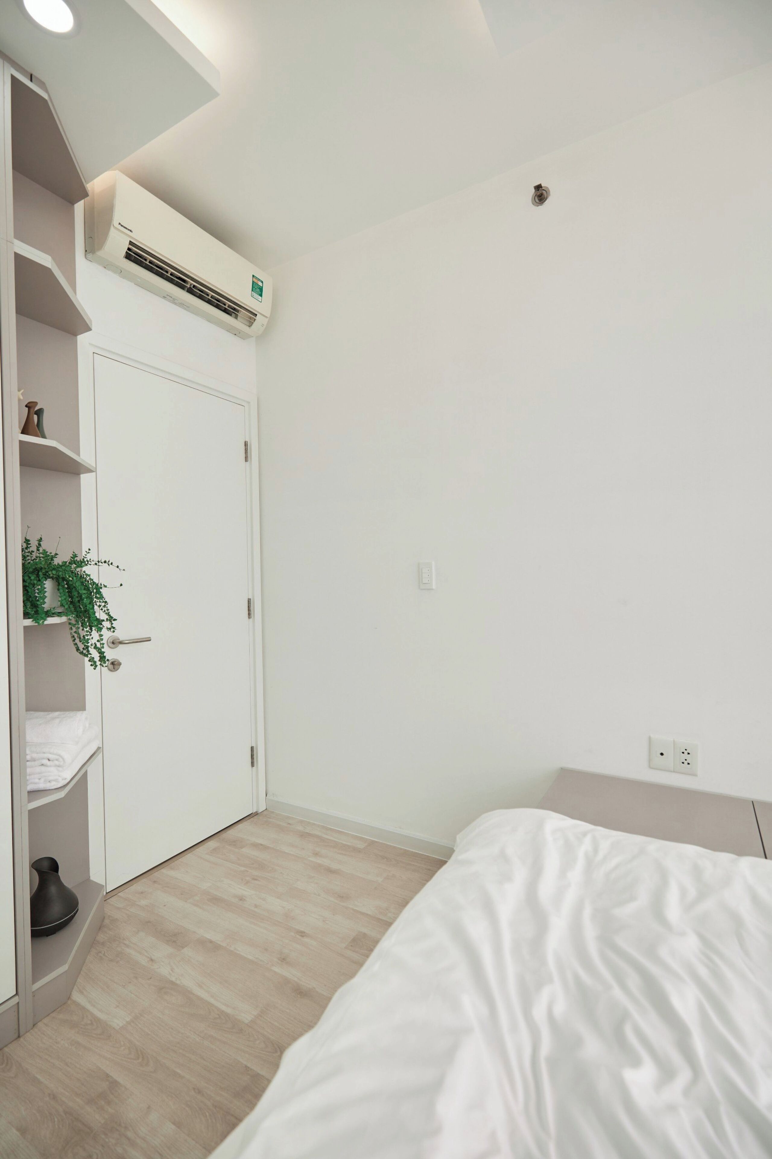 A bedroom with a bed, a wardrobe, a shelf with a plant, and an air conditioning unit on the wall.