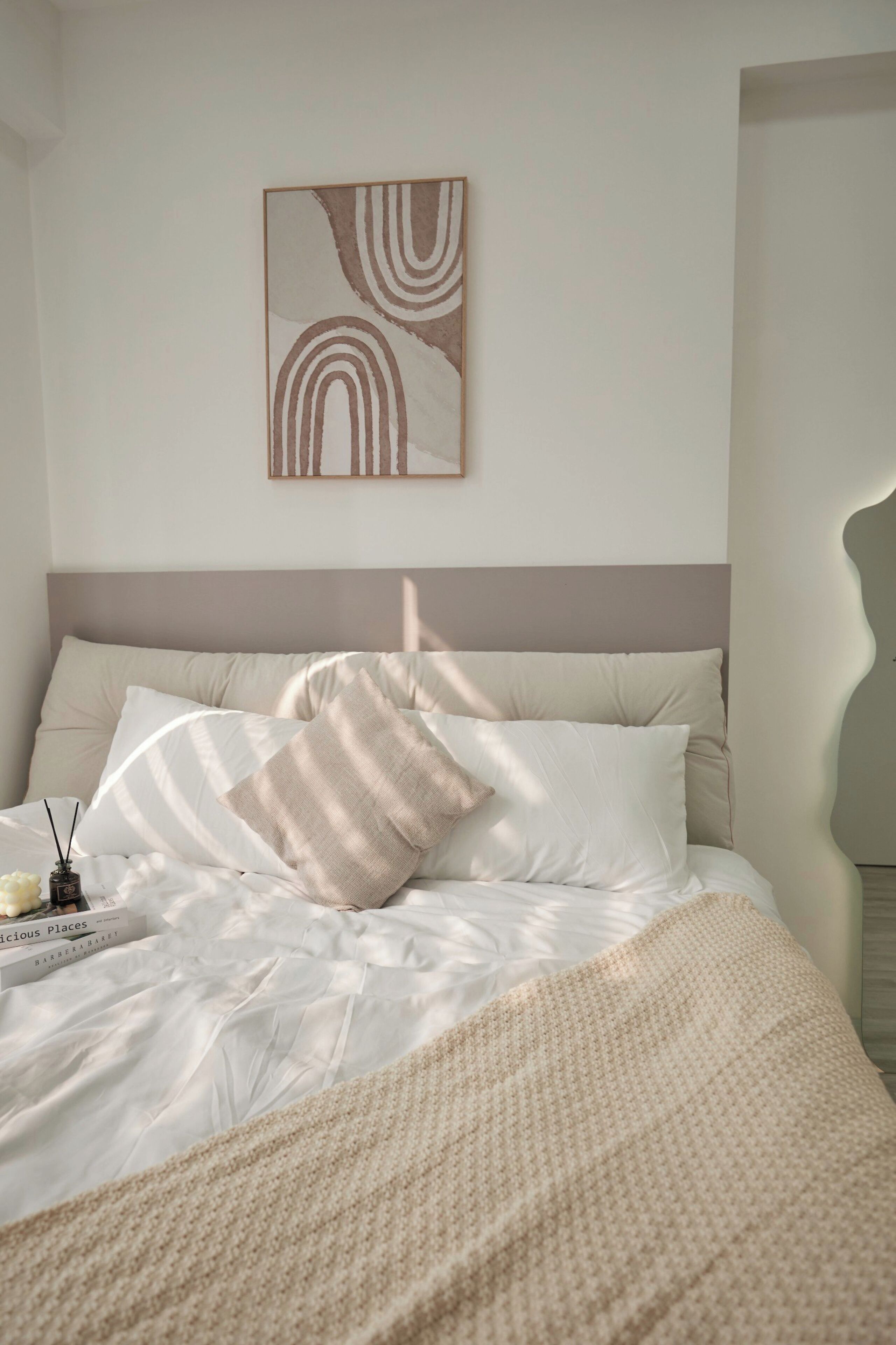 A neatly made bed with a beige blanket and pillows, a framed abstract artwork on the wall, and a bedside table with a book and a candle.