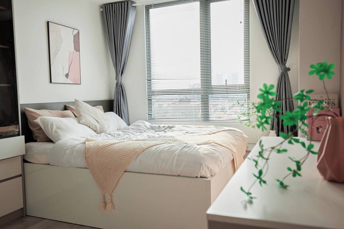 Ein modernes Schlafzimmer mit Bett, weißen Wänden, einem Fenster mit Jalousien und einem Nachttisch mit Pflanze.
