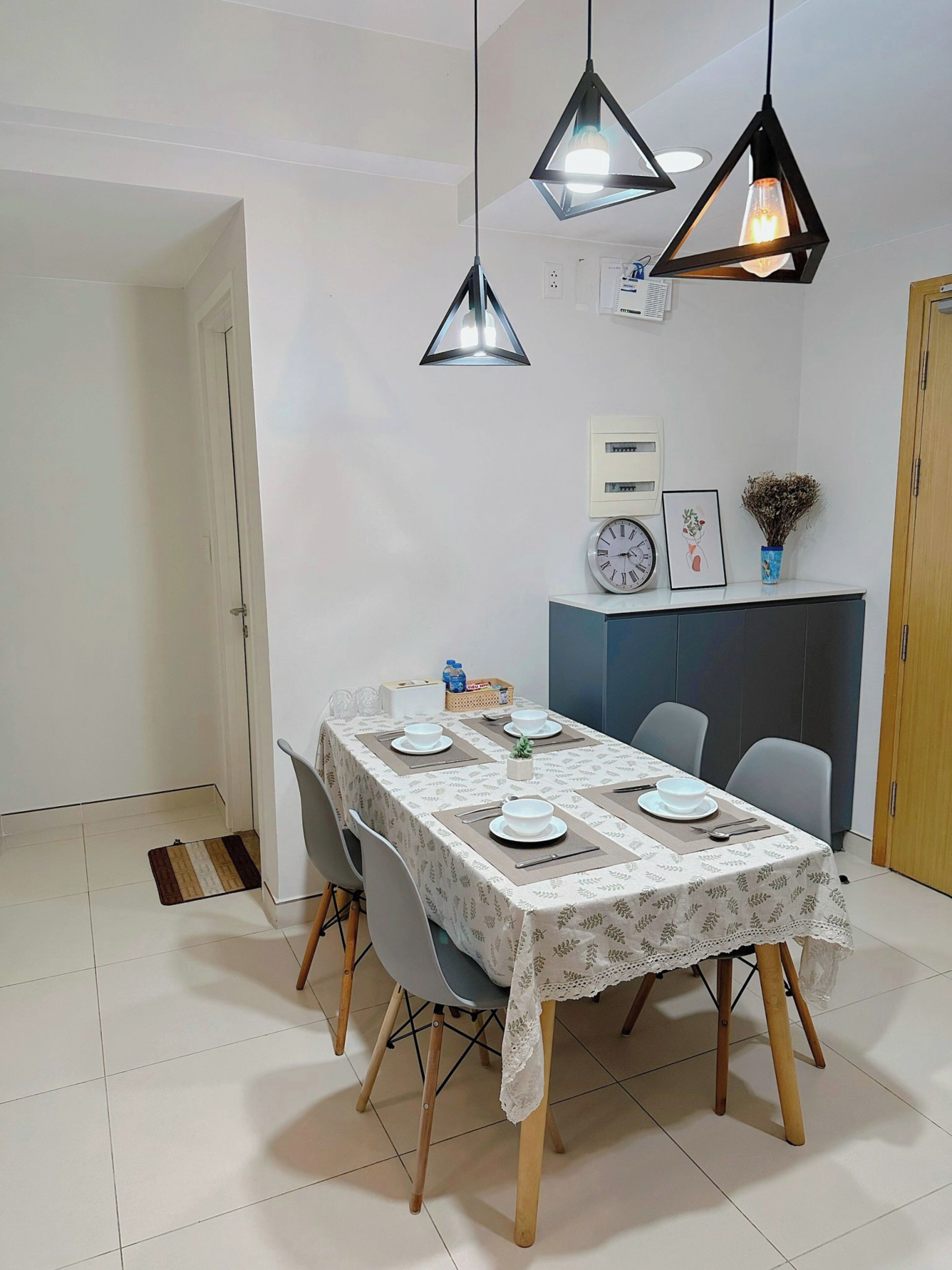 Un comedor con una mesa dispuesta para cuatro personas, dos lámparas colgantes, un reloj y una pequeña planta en la pared.