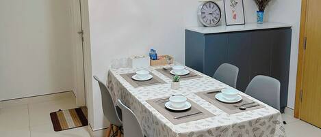 Un coin repas avec une table dressée pour quatre personnes, deux lampes suspendues, une horloge et une petite plante fixée au mur.